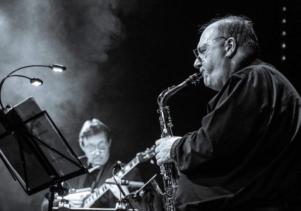 Black and white photograph of a saxophonist performing in close-up. An older male musician wearing glasses plays an alto or soprano saxophone, captured in profile against a dark background with atmospheric stage lighting. A keyboard player is visible out of focus in the background.
