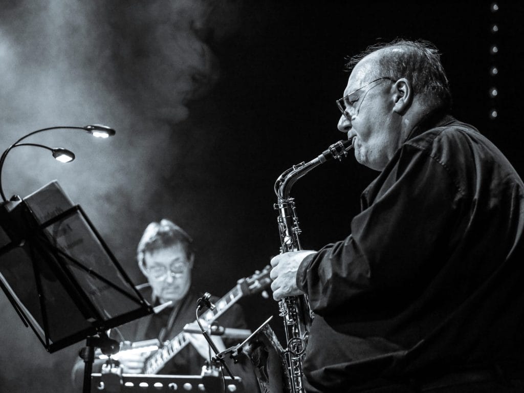 Black and white photograph of a saxophonist performing in close-up. An older male musician wearing glasses plays an alto or soprano saxophone, captured in profile against a dark background with atmospheric stage lighting. A keyboard player is visible out of focus in the background.
