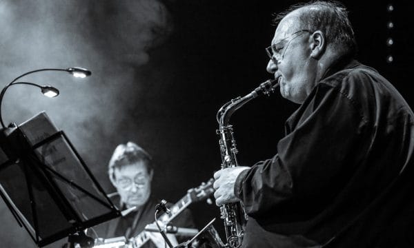 Black and white photograph of a saxophonist performing in close-up. An older male musician wearing glasses plays an alto or soprano saxophone, captured in profile against a dark background with atmospheric stage lighting. A keyboard player is visible out of focus in the background.