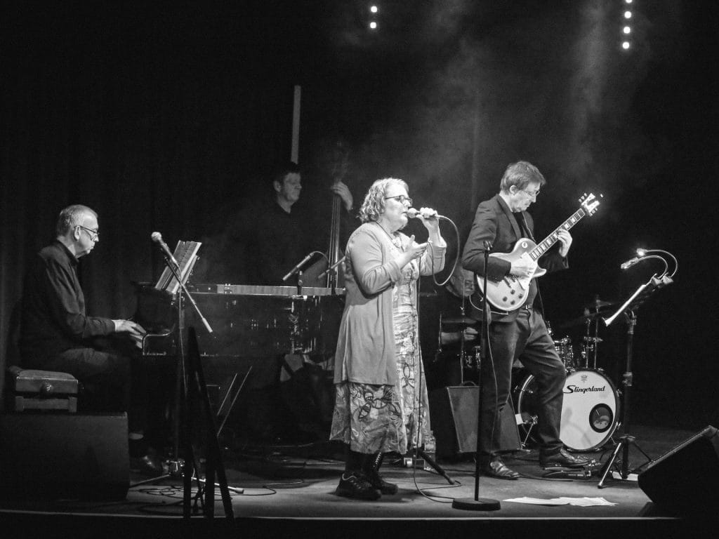 Black and white photograph of a jazz ensemble performing on stage. A female vocalist in glasses stands at centre microphone wearing a cardigan and patterned dress, flanked by a guitarist on the right playing a Les Paul-style electric guitar and a keyboard player on the left. A double bass player and drummer are visible in the background, with atmospheric stage lighting and smoke creating dramatic effect