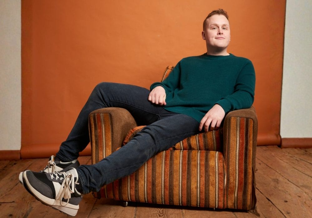 A young man wearing a dark blue jumper and dark coloured jeans sitting casually on a brown stripy arm chair
