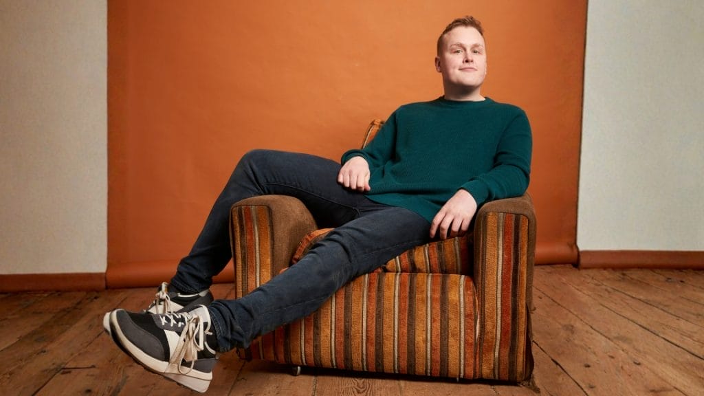 A young man wearing a dark blue jumper and dark coloured jeans sitting casually on a brown stripy arm chair