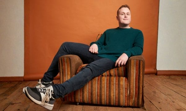 A young man wearing a dark blue jumper and dark coloured jeans sitting casually on a brown stripy arm chair