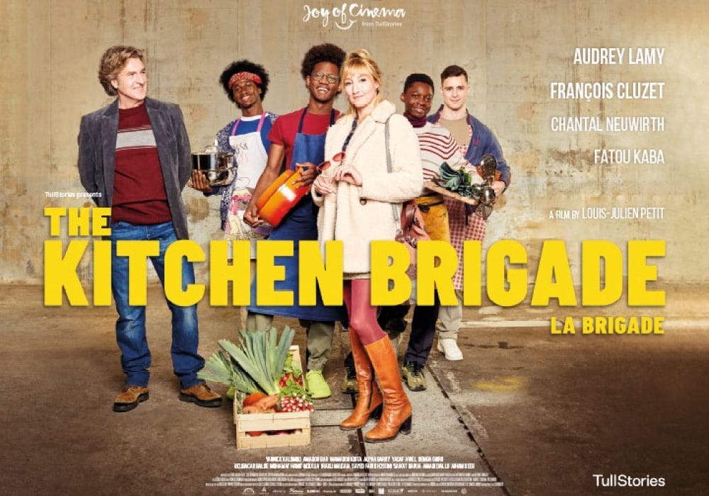 A group of people standing together holding cooking pans. There is a crate of fruit and vegetables on the floor