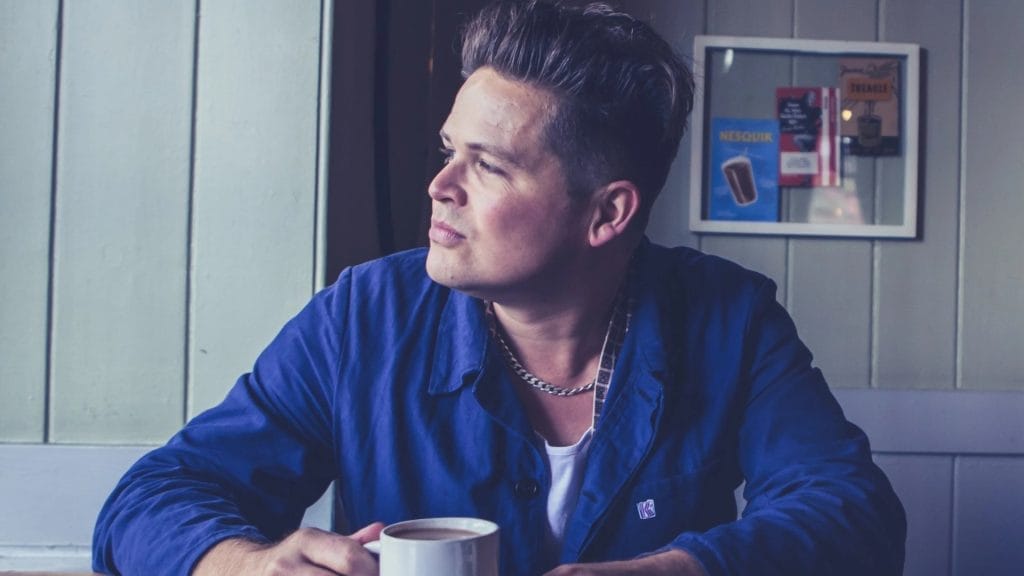 A man with quiffed hair and wearing a blue shirt sitting at table holding a cup of tea whilst looking out of a window