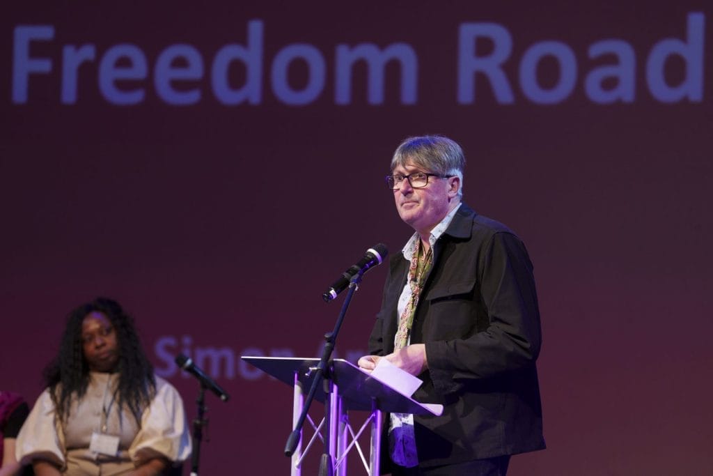 A man speaks at a podium with a microphone, set against a purple backdrop displaying "Freedom Road." A woman sits attentively in the background. The atmosphere is formal.