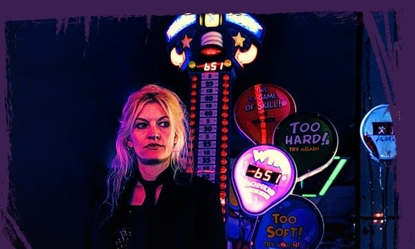 A blonde haired woman dressed in black standing in front of Las Vegas style neon lighting