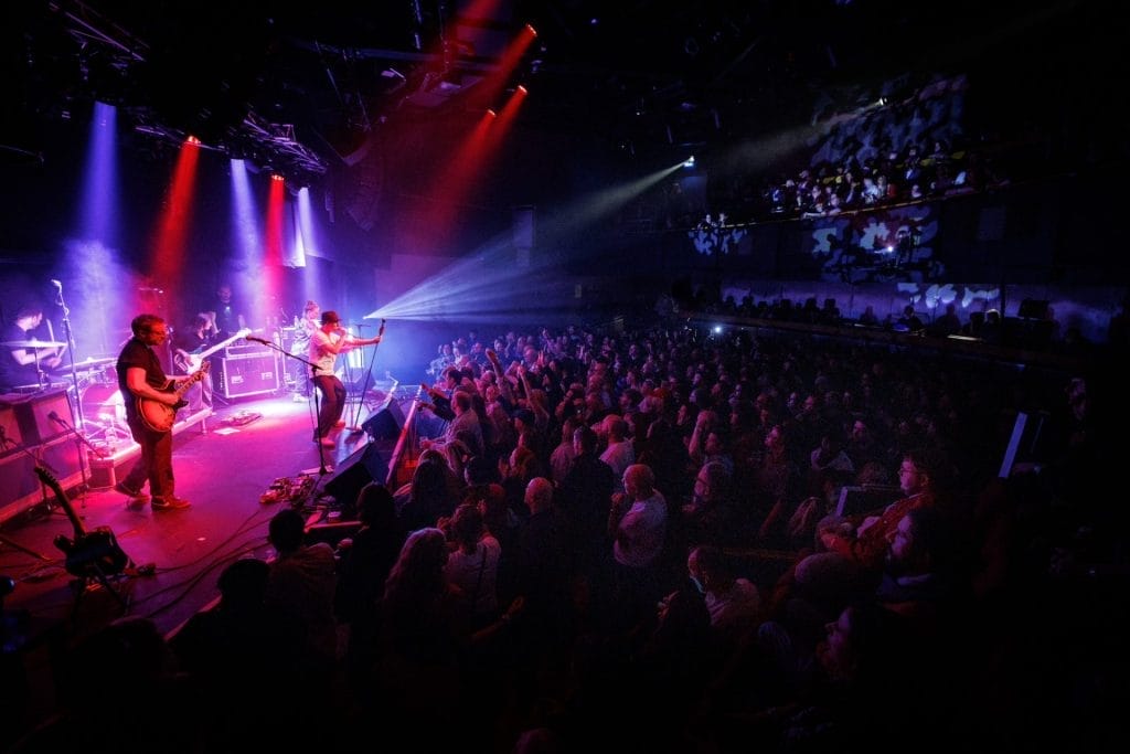 A lively band plays on stage, surrounded by bright lights that enhance the dynamic performance for the crowd.