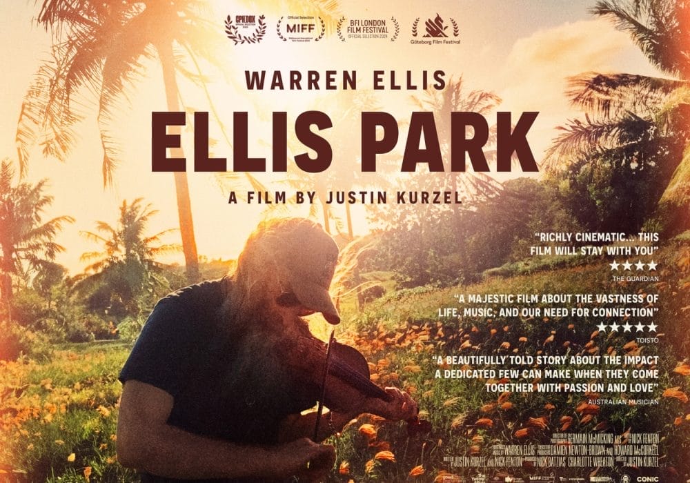 A bearded man wearing a baseball cap and sunglasses playing a violin in a tropical field