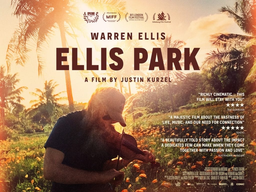 A bearded man wearing a baseball cap and sunglasses playing a violin in a tropical field