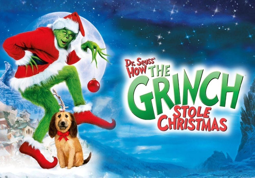 A human sized furry green creature wearing a red Santa outfit holding a red bauble in his hand. There is a brown long haired dog sitting at his feet. They are both standing in a snow covered landscape