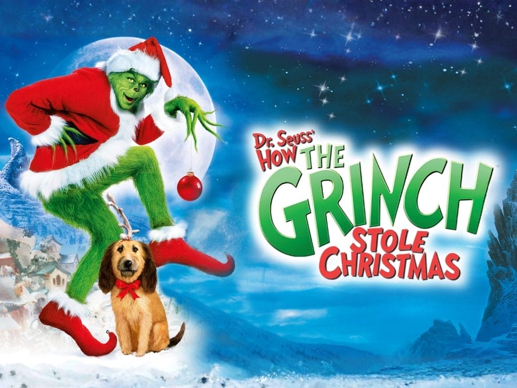 A human sized furry green creature wearing a red Santa outfit holding a red bauble in his hand. There is a brown long haired dog sitting at his feet. They are both standing in a snow covered landscape