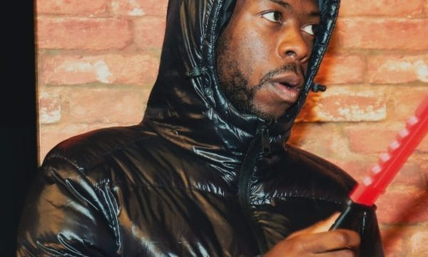 A person in a shiny black jacket holding a red knife poses in front of a brick wall, highlighting a pivotal moment.